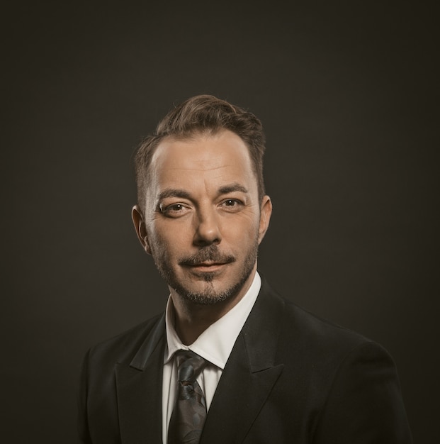 Apuesto hombre de negocios positivo mira a la cámara. Vista frontal del alegre hombre caucásico en elegante traje negro sonriendo aislado sobre fondo gris. Imagen tonificada