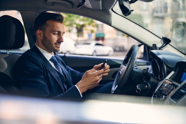 Apuesto hombre de negocios en ropa formal pasar tiempo en auto mientras mira en el teléfono inteligente