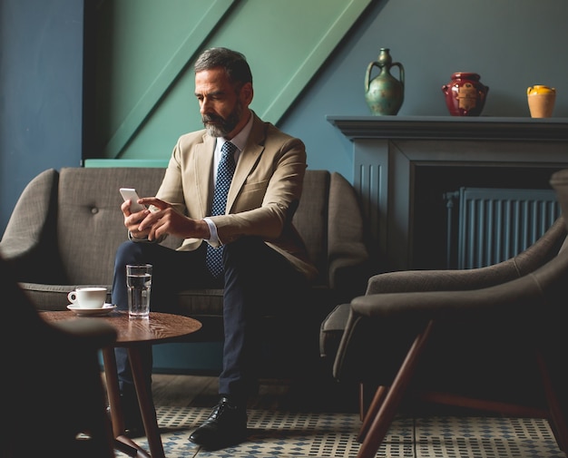 Apuesto hombre de negocios senior tomando café y usando el teléfono móvil en el vestíbulo