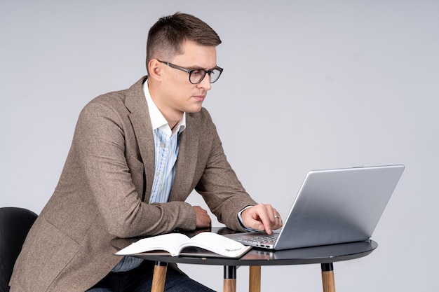 Apuesto hombre de negocios sentado en la mesa y trabajando en la computadora portátil Hombre joven en traje está sentado y mirando la pantalla Retrato de cuerpo entero