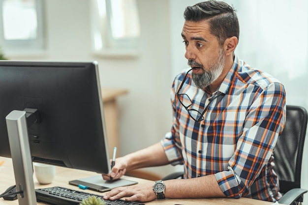 Un apuesto hombre de negocios serio y pensativo está trabajando en una computadora en la oficina. Está sorprendido de que algo se muestre en la computadora.