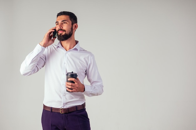 Apuesto hombre de negocios de Sheikh árabe con barba negra en una camisa blanca y pantalones negros sosteniendo una taza de papel de plástico y bebiendo café usando un teléfono inteligente en una foto de estudio de fondo blanco