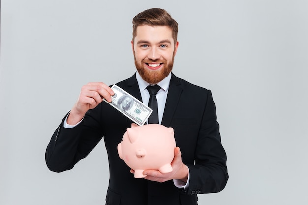 Apuesto hombre de negocios sonriente en traje poniendo dinero en la hucha