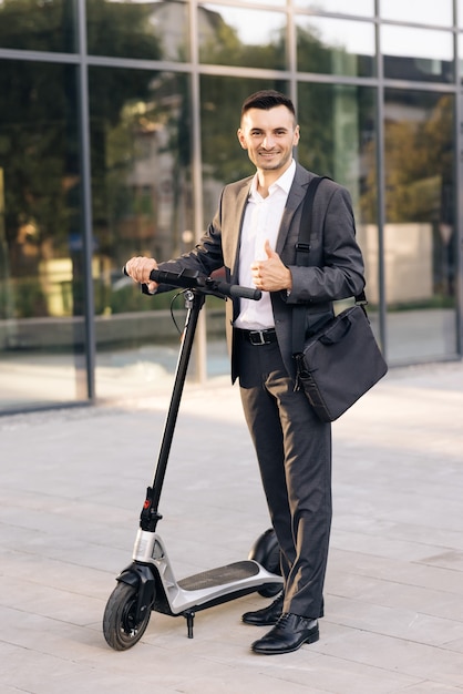 Apuesto hombre de negocios con su scooter eléctrico frente a la construcción de negocios joven empresario