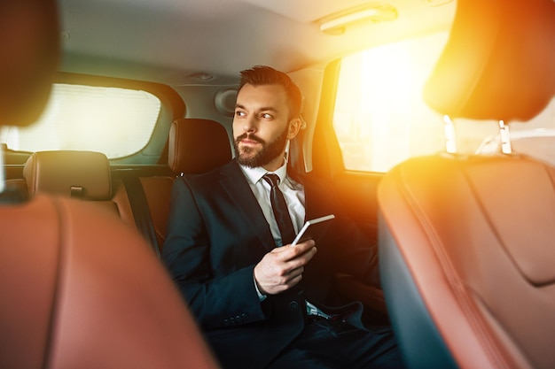 Apuesto hombre de negocios con teléfono en la mano sentado en el asiento trasero del auto mirando hacia otro lado