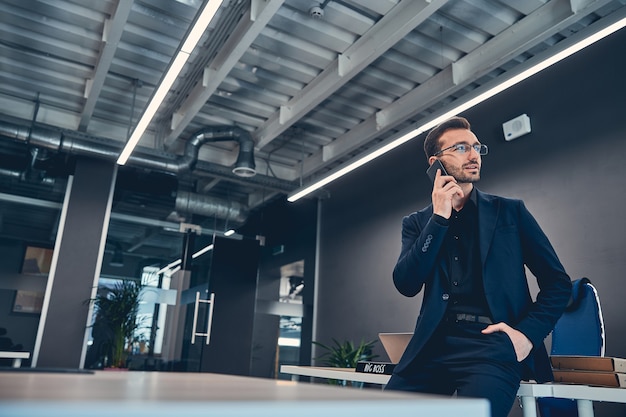 Apuesto hombre de negocios en traje y anteojos hablando por teléfono en el lugar de trabajo