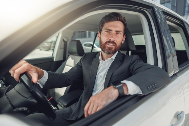 Apuesto hombre de negocios en traje gris está montando detrás del volante del coche