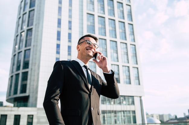 Apuesto hombre de negocios con traje negro completo habla en su teléfono inteligente en el fondo del paisaje urbano
