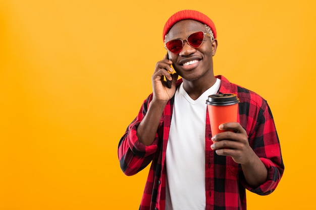 Apuesto hombre negro americano hablando por teléfono mientras sostiene un vaso de café para llevar sobre fondo amarillo studio