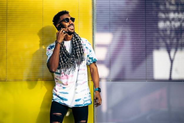 Un apuesto hombre negro sonriente apoyado contra una pared de vidrio púrpura y amarillo usando su teléfono inteligente