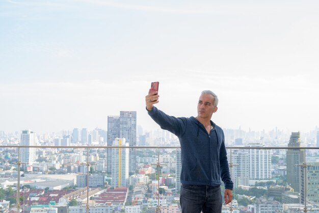 Apuesto hombre persa con cabello gris tomando selfie contra vista de la ciudad