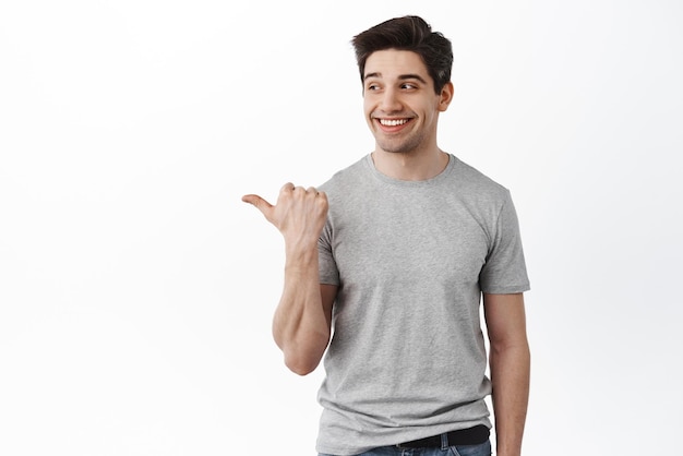 Un apuesto hombre sano adulto señalando y mirando a un lado el texto promocional que muestra una pancarta y sonriendo de pie en una camiseta contra el fondo blanco