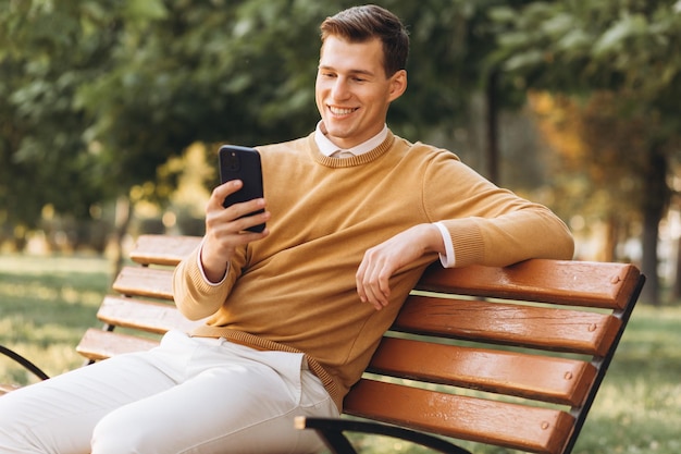 Un apuesto hombre sonriente moderno con ropa amarilla y blanca hablando por teléfono sentado en un banco del parque al atardecer