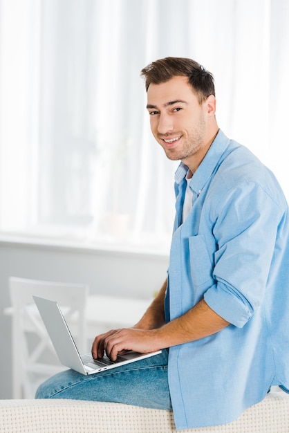 Un apuesto hombre sonriente sentado mirando la cámara y usando una laptop en casa con espacio para copiar