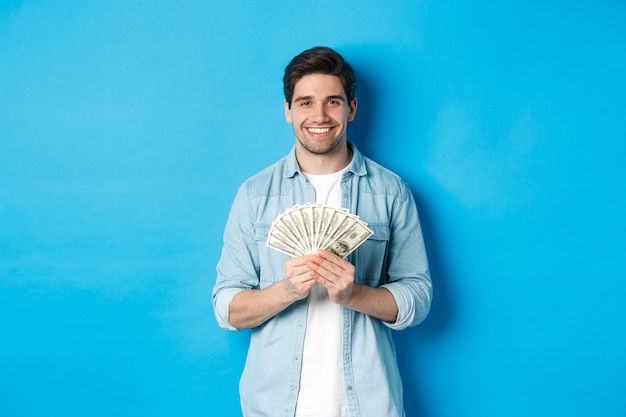 Apuesto hombre sonriente sosteniendo dinero, concepto de finanzas y banca, de pie sobre la pared azul