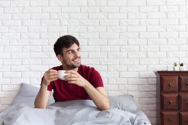 Apuesto hombre sonriente sosteniendo una taza de café y sentado en la cama