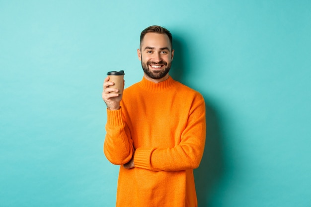 Apuesto hombre sonriente tomando un descanso, tomando café de comida para llevar, de pie sobre fondo azul.