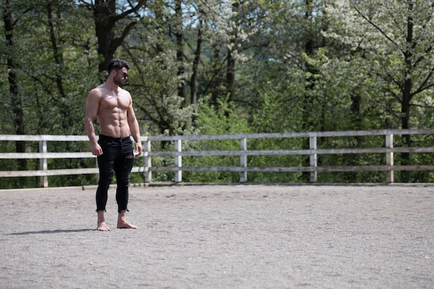 Apuesto hombre vaquero de pie fuerte posando en el rancho al aire libre usando jeans negros y sombrero de vaquero