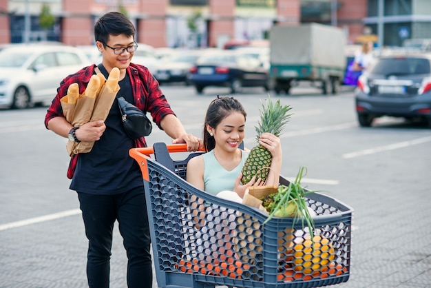 Apuesto hombre vietnamita sostiene bolsas de papel con comida empujando frente a él carrito de compras con su hermosa novia feliz dentro de él.