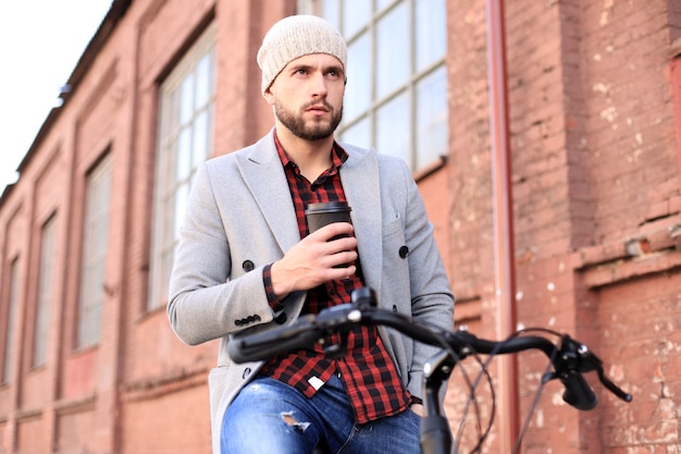 Apuesto joven con abrigo gris y sombrero y andar en bicicleta por la calle tomando café.