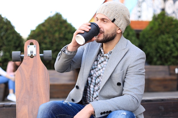 Apuesto joven con abrigo gris y sombrero, descansando, sentado con longboard tomando café. Concepto de skate urbano.