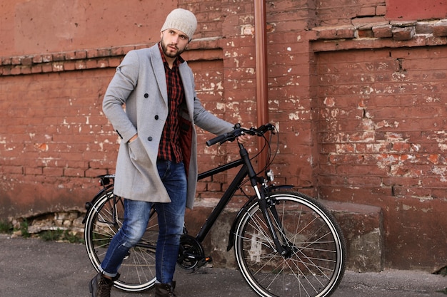 Apuesto joven con abrigo gris y sombrero empujando su bicicleta por una calle de la ciudad.