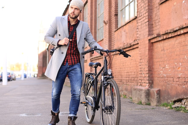 Apuesto joven con abrigo gris y sombrero empujando su bicicleta por una calle de la ciudad.