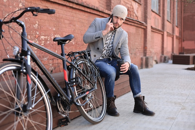 Apuesto joven con abrigo gris y sombrero hablando por teléfono móvil y sonriendo mientras está sentado cerca de su bicicleta al aire libre.