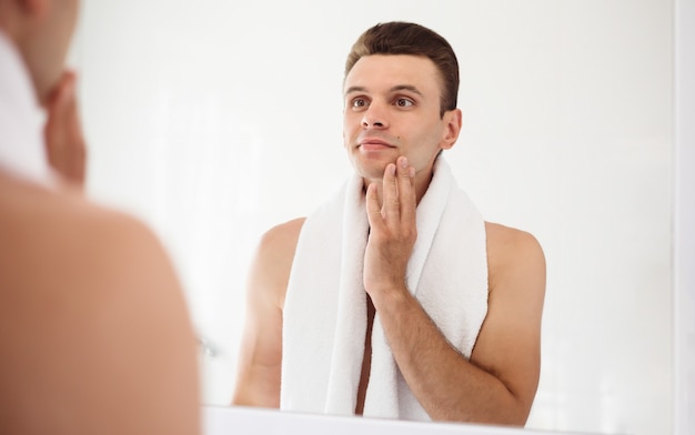 Apuesto joven afeitándose la barba en el baño. Retrato de un elegante hombre barbudo desnudo examinando su rostro en el espejo de la casa.