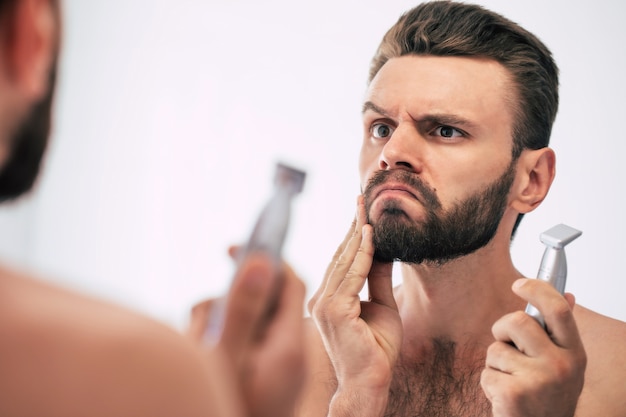 Apuesto joven afeitándose la barba en el baño. Retrato de un elegante hombre barbudo desnudo examinando su rostro en el espejo de la casa.