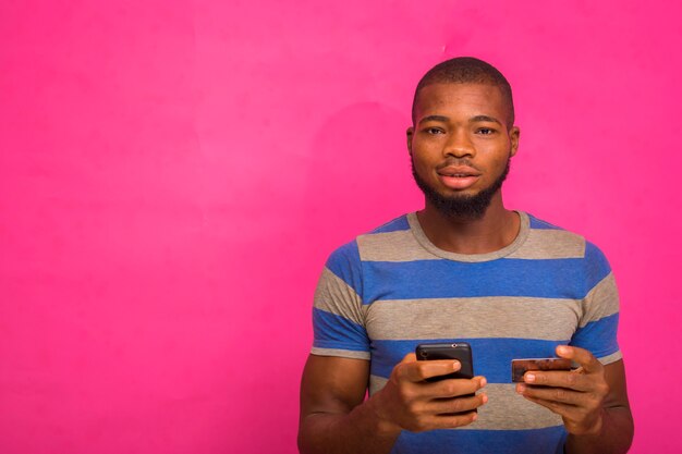 Apuesto joven africano aislado sobre fondo rosa sosteniendo su tarjeta de crédito y teléfono inteligente.