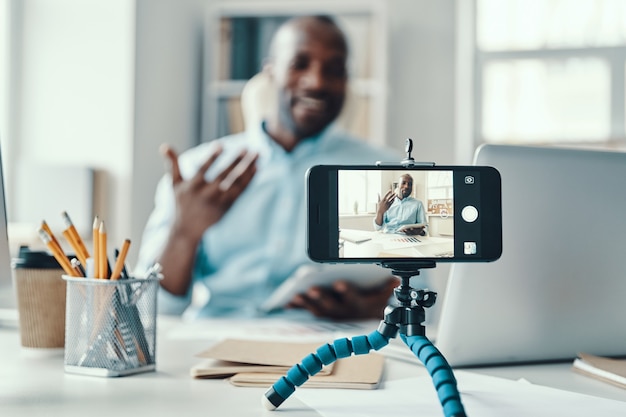 Apuesto joven africano en camisa diciendo algo y sonriendo mientras hace videos de redes sociales