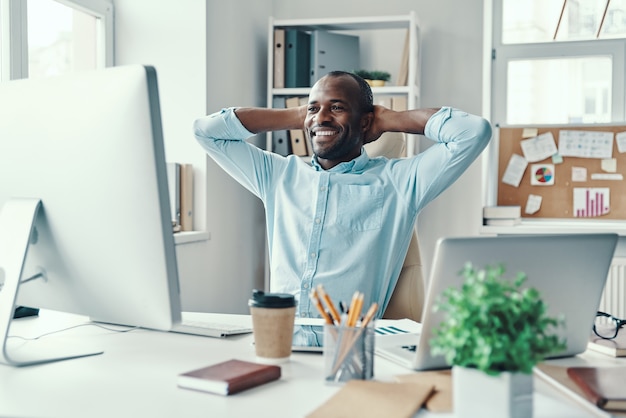 Apuesto joven africano en camisa manteniendo las manos detrás de la cabeza y sonriendo mientras trabaja en la oficina