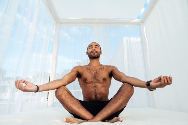 Apuesto joven africano en pantalones cortos sentado y meditando en la cama de la playa