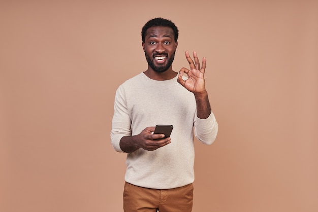 Apuesto joven africano en ropa casual con teléfono inteligente y mostrando un gesto bien