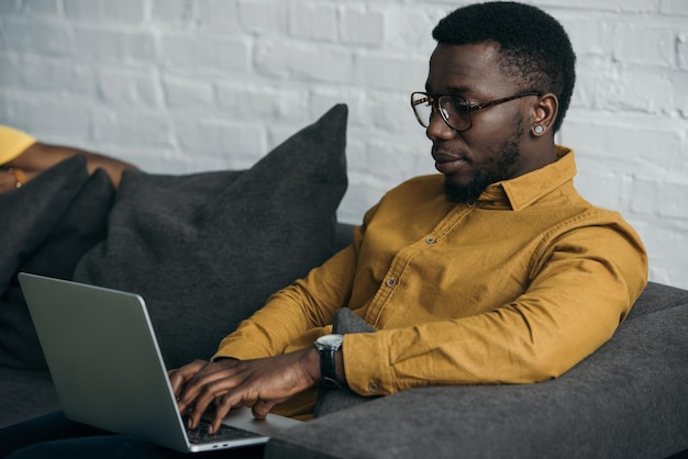 Apuesto joven afroamericano en anteojos sentado en el sofá y usando una computadora portátil