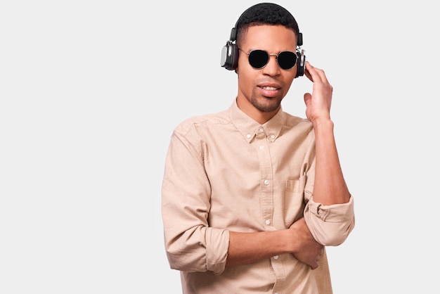 Apuesto joven afroamericano con auriculares escuchando la música favorita Estudiante afro con gafas de sol y ropa informal posando sobre fondo blanco de estudio