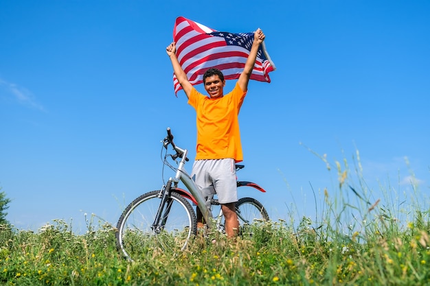 Apuesto joven afroamericano sosteniendo y ondeando la bandera de Estados Unidos y de pie con bicicleta en la pradera de verano contra el fondo de cielo azul en la mañana