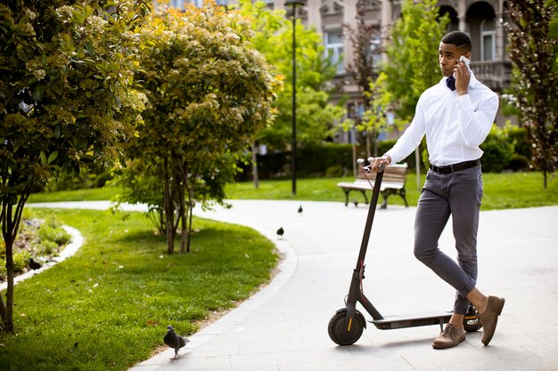 Apuesto joven afroamericano mediante teléfono móvil mientras está de pie por scooter eléctrico en una calle