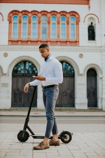 Apuesto joven afroamericano mediante teléfono móvil mientras está de pie con scooter eléctrico en una calle