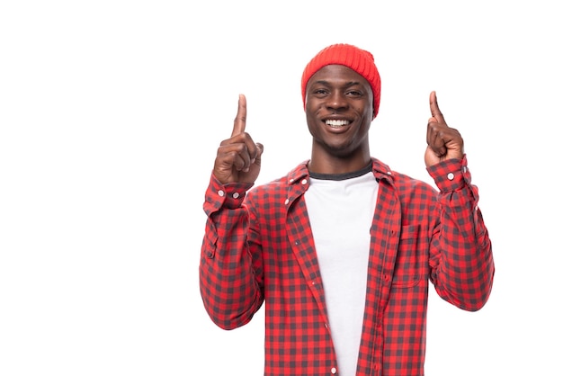 Apuesto joven americano en gorra roja y camisa a cuadros apuntando con el dedo sobre fondo blanco con