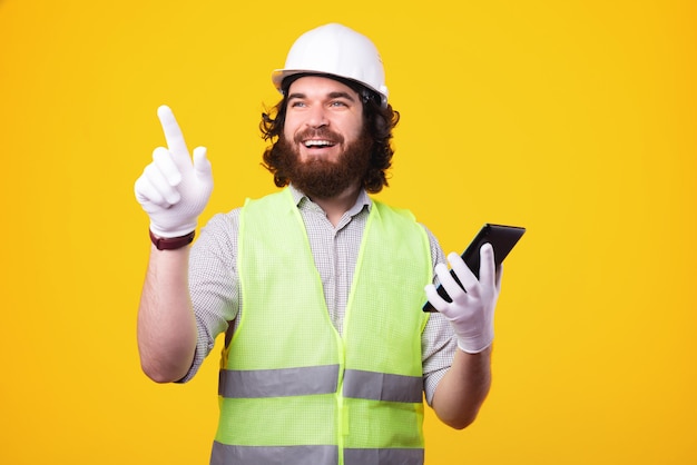 Apuesto joven arquitecto hombre sujetando la tableta y apuntando hacia fuera sobre fondo amarillo