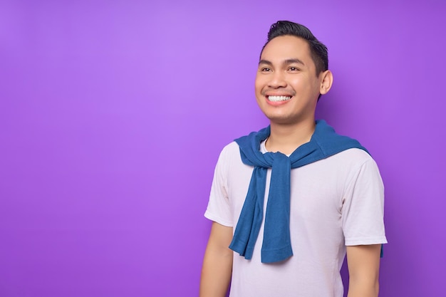 Apuesto joven asiático de 20 años con camiseta blanca mirando a un lado en el área de trabajo con una sonrisa dentuda aislada en un fondo morado Concepto de estilo de vida de la gente