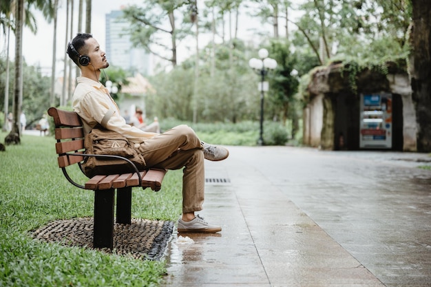 Un apuesto joven asiático escuchando y disfrutando de música conmovedora con auriculares y ojos cerrados sentado en un banco en un parque con una bolsa durante la mañana y la noche