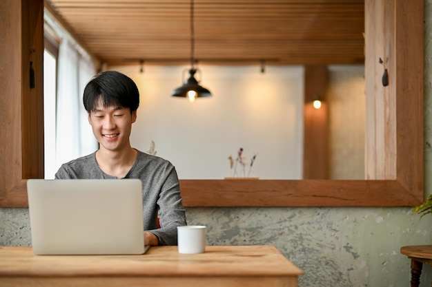 Un apuesto joven asiático con ropa informal usando su laptop trabajando a distancia en la cafetería