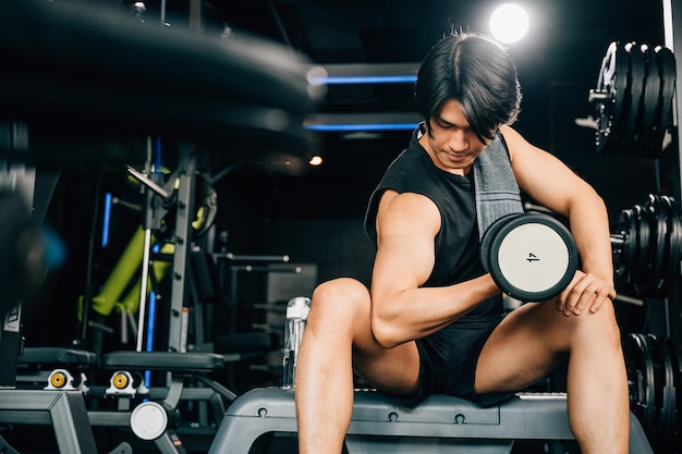 Un apuesto joven asiático sentado en un banco de ejercicios sosteniendo una pesa pesada mostrando su musculoso brazo y bíceps y su dedicación al espacio vacío del fitness