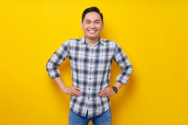 Un apuesto joven asiático sonriendo con un pantalones a cuadros de pie posando con una mano en la cintura mientras mira la cámara aislada con confianza en el concepto de estilo de vida de la gente de fondo amarillo