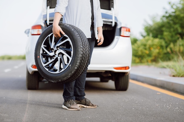 Foto apuesto joven asiático sujetando la rueda de repuesto para cambiar la rueda desinflada en la carretera