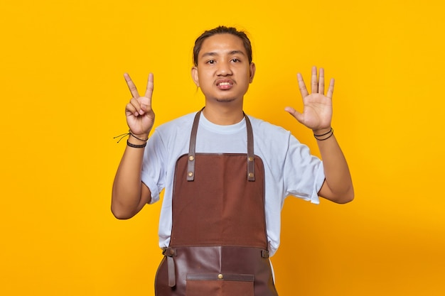 Apuesto joven asiático vistiendo delantal apuntando con el dedo número siete con expresión de enojo