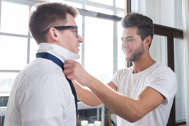 Apuesto joven atando la corbata de sus novios mientras pasan tiempo juntos y disfrutan del día.